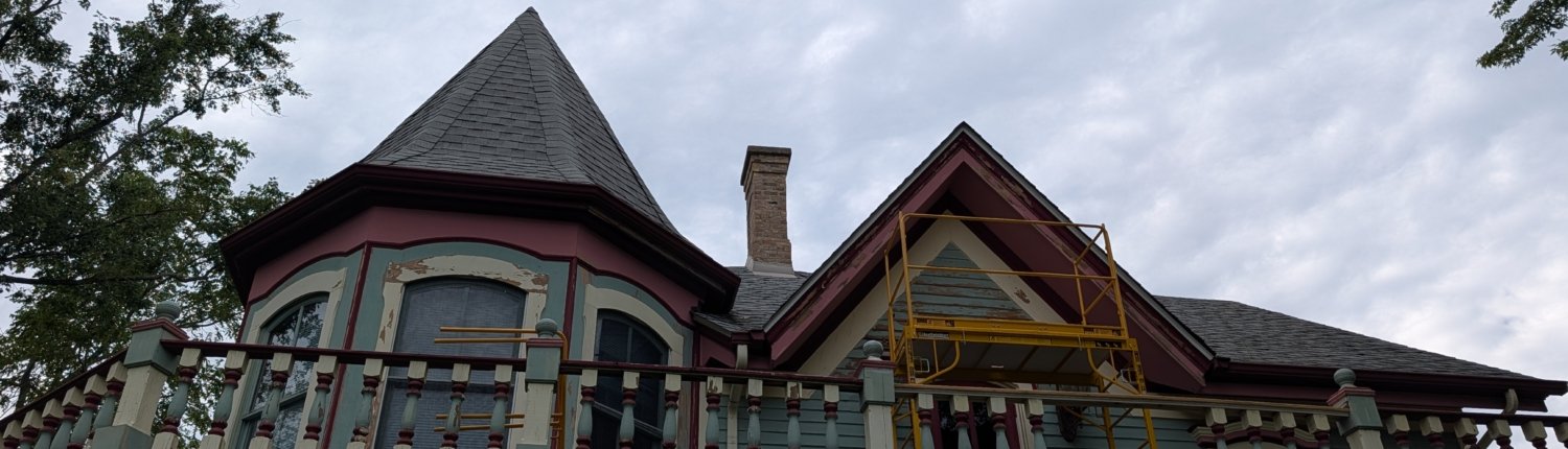 Victorian house undergoing exterior repairs A low-angle view looking up at the upper story and roofline of a Victorian house undergoing exterior repairs, featuring a conical turret roof, balustraded balcony, and yellow scaffolding against a cloudy sky.
