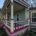 A side-angle view of a colorful Victorian wrap-around porch featuring painted balusters, cream columns, light blue siding, and a bay window framed in maroon and cream trim