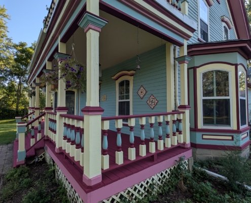 A side-angle view of a colorful Victorian wrap-around porch featuring painted balusters, cream columns, light blue siding, and a bay window framed in maroon and cream trim